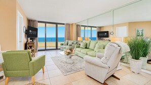 En rummelig stue med en grøn sofa, en beige lænestol og et sofabord. Der er et stort vindue med udsigt over havet.