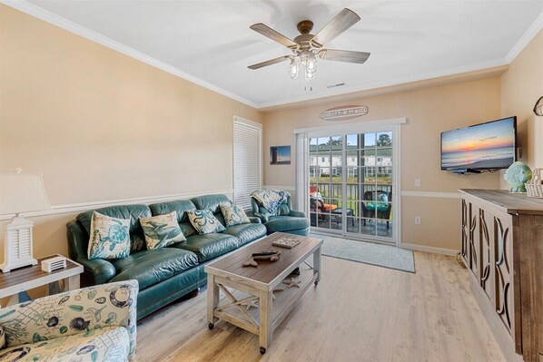 A living room with a green sofa, a wooden coffee table, a ceiling fan, and a TV mounted on the wall.