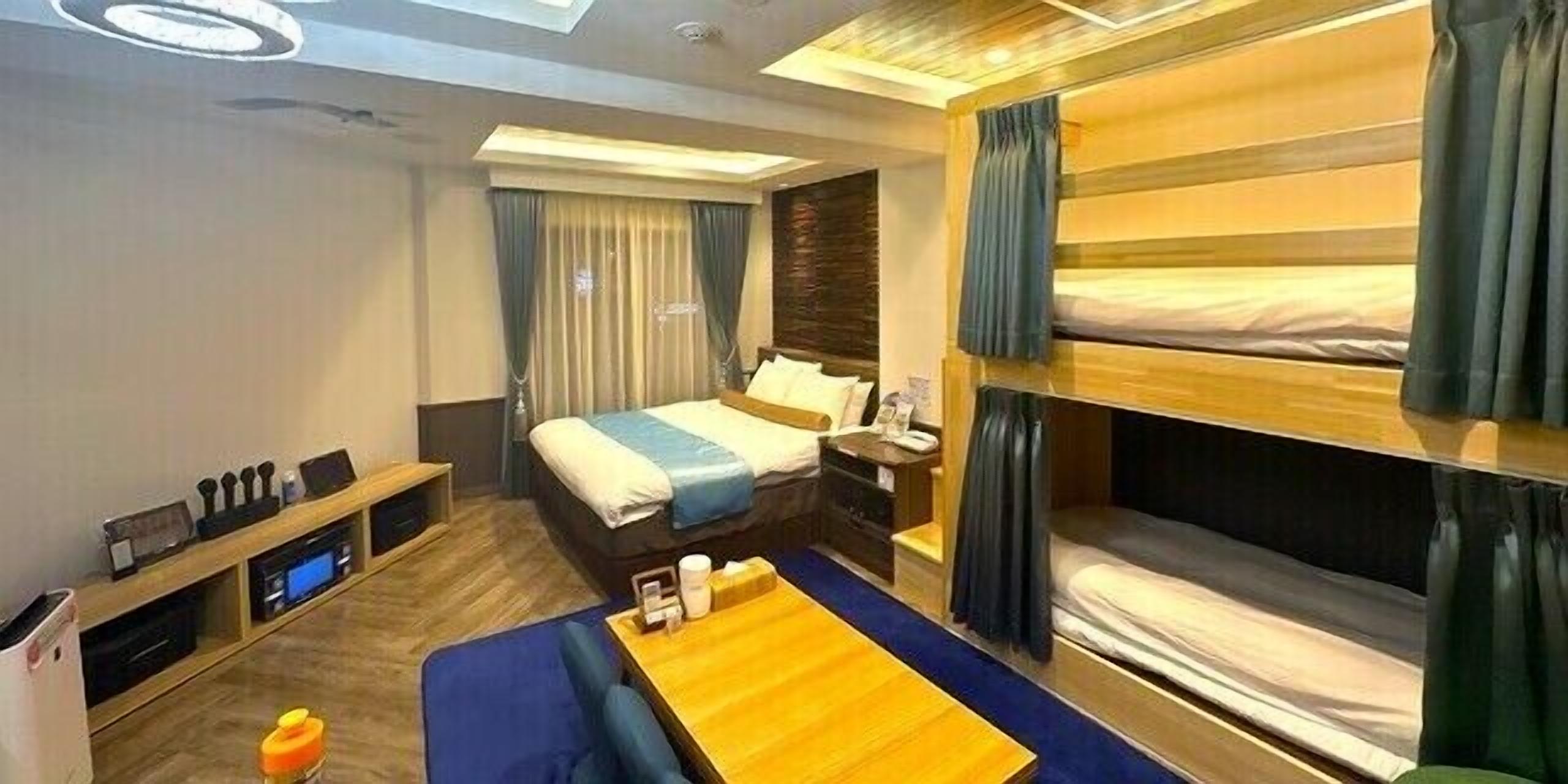 A hotel room with a bunk bed, a desk with a computer, a bedside table, and a large window with curtains.