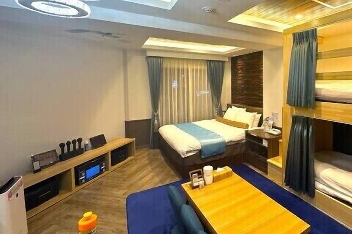 A hotel room with a bunk bed, a desk with a computer, a bedside table, and a large window with curtains.