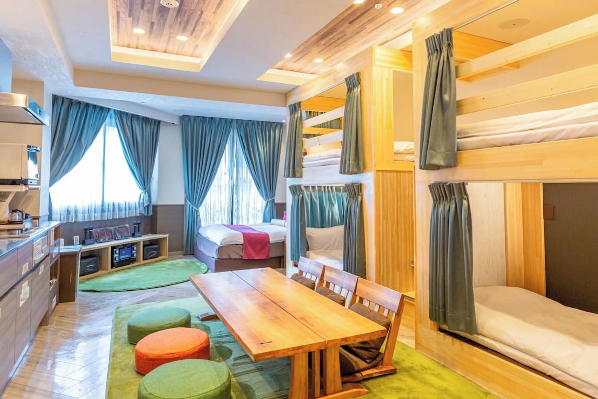 A compact hotel room with bunk beds, a dining table, a kitchenette, and a large window with curtains.