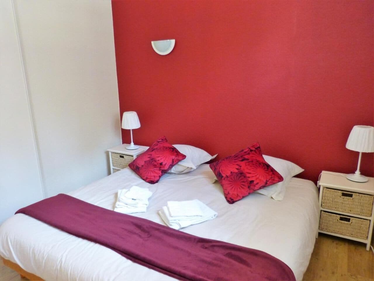 A neatly made bed with a red blanket, two red pillows, and white linens. There are two bedside tables with lamps and a wall-mounted light.