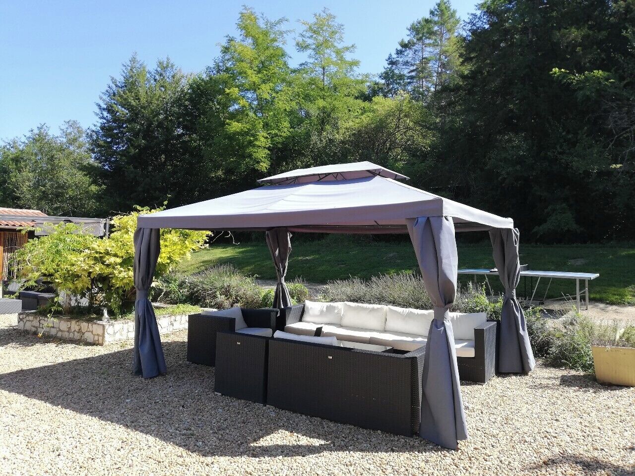 A covered outdoor seating area with a table and chairs, surrounded by gravel and greenery.