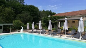 A swimming pool with lounge chairs and umbrellas, surrounded by greenery and a stone building. - Chambres d'hôtes La Vergne (Saint-Martin-des-Combes)