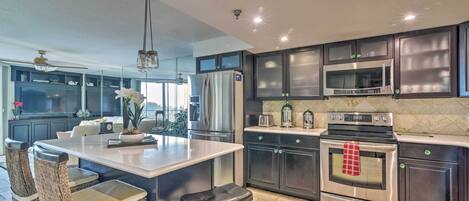 A modern kitchen with dark cabinets, stainless steel appliances, a large island with bar stools, and a view to the outdoors.
