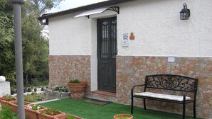 A small building with a stone and plaster exterior, a black door, a bench, and potted plants. - The farther you are from the world, the more beautiful the world seems (Noto (SR))