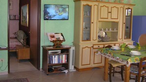 A dining area with a table set for four, a television mounted on the wall, and a bookshelf with books and a DVD player. - The farther you are from the world, the more beautiful the world seems (Noto (SR))
