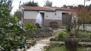 A rustic building with a stone wall, a water feature, and a bench. - The farther you are from the world, the more beautiful the world seems (Noto (SR))