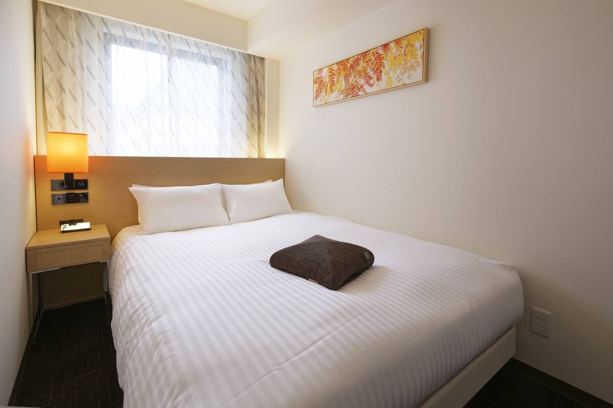 Ein Hotelzimmer mit einem Bett, einem Nachttisch, einer Lampe, einem Fenster mit Vorhängen und einem Wandbild.