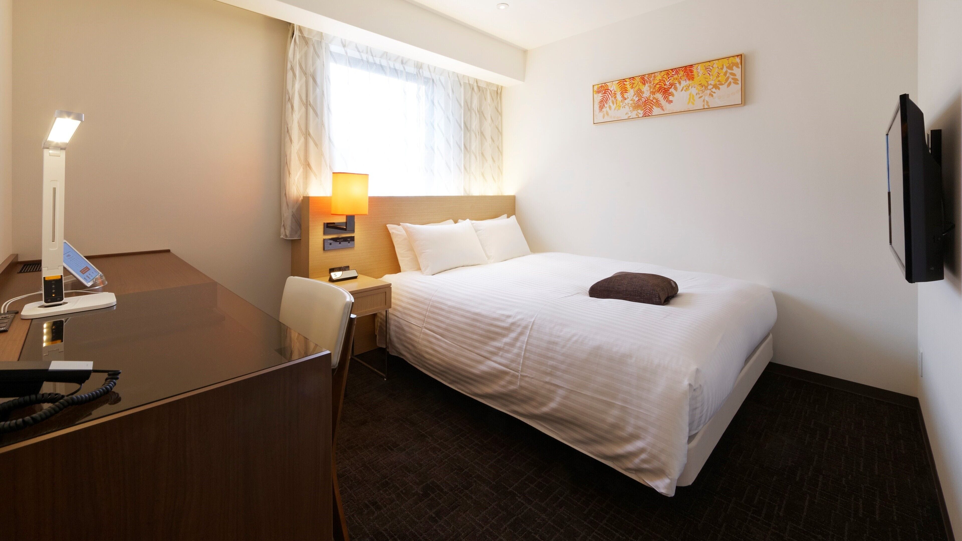 Ein Hotelzimmer mit Bett, Schreibtisch, Stuhl, Lampe und Kunstwerk an der Wand.