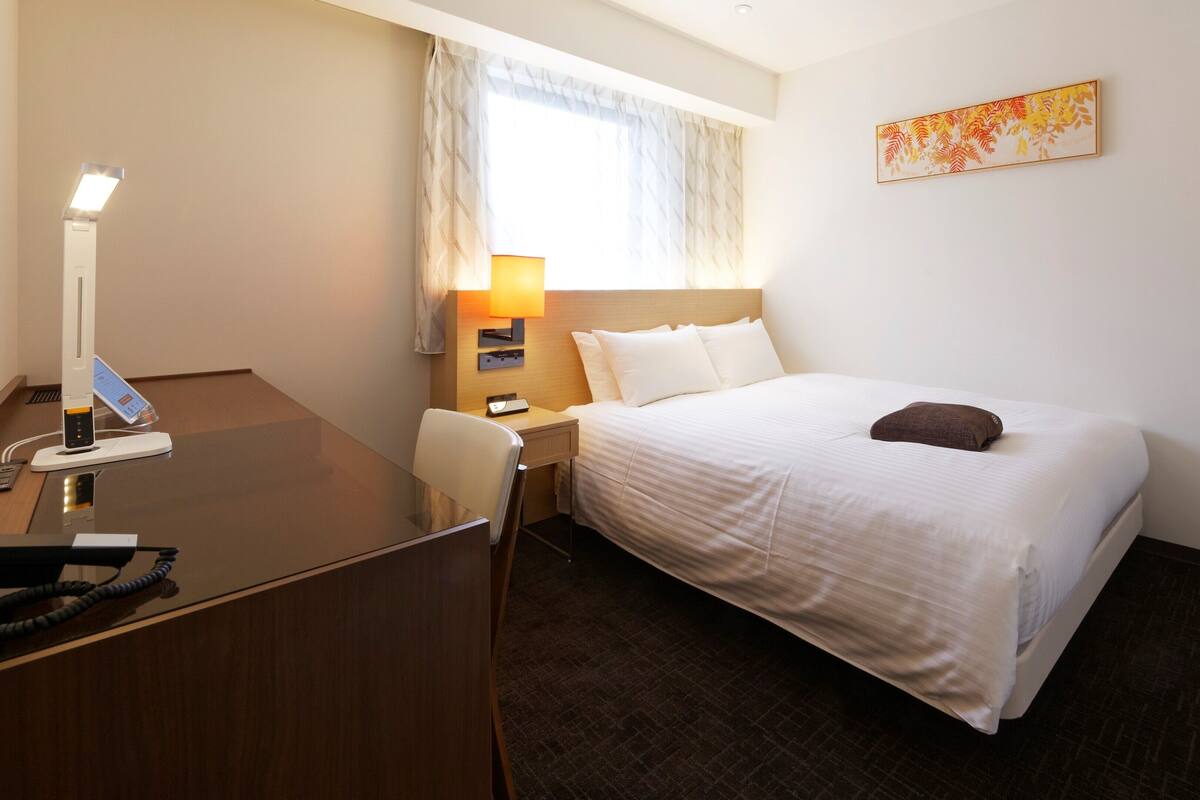 Ein Hotelzimmer mit Bett, Schreibtisch, Stuhl, Lampe und Kunstwerk an der Wand.