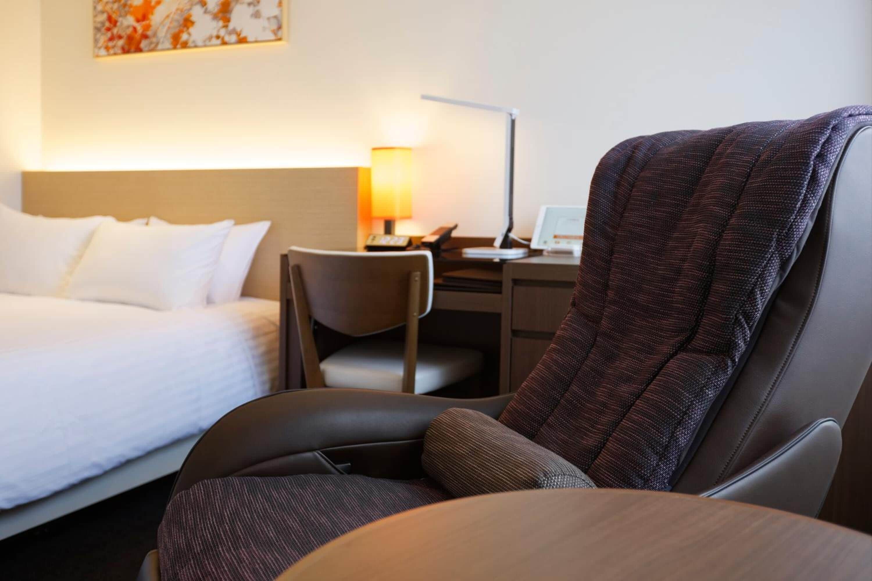 Una habitación de hotel con una cama, un escritorio con lámpara, una silla y un sillón grande.