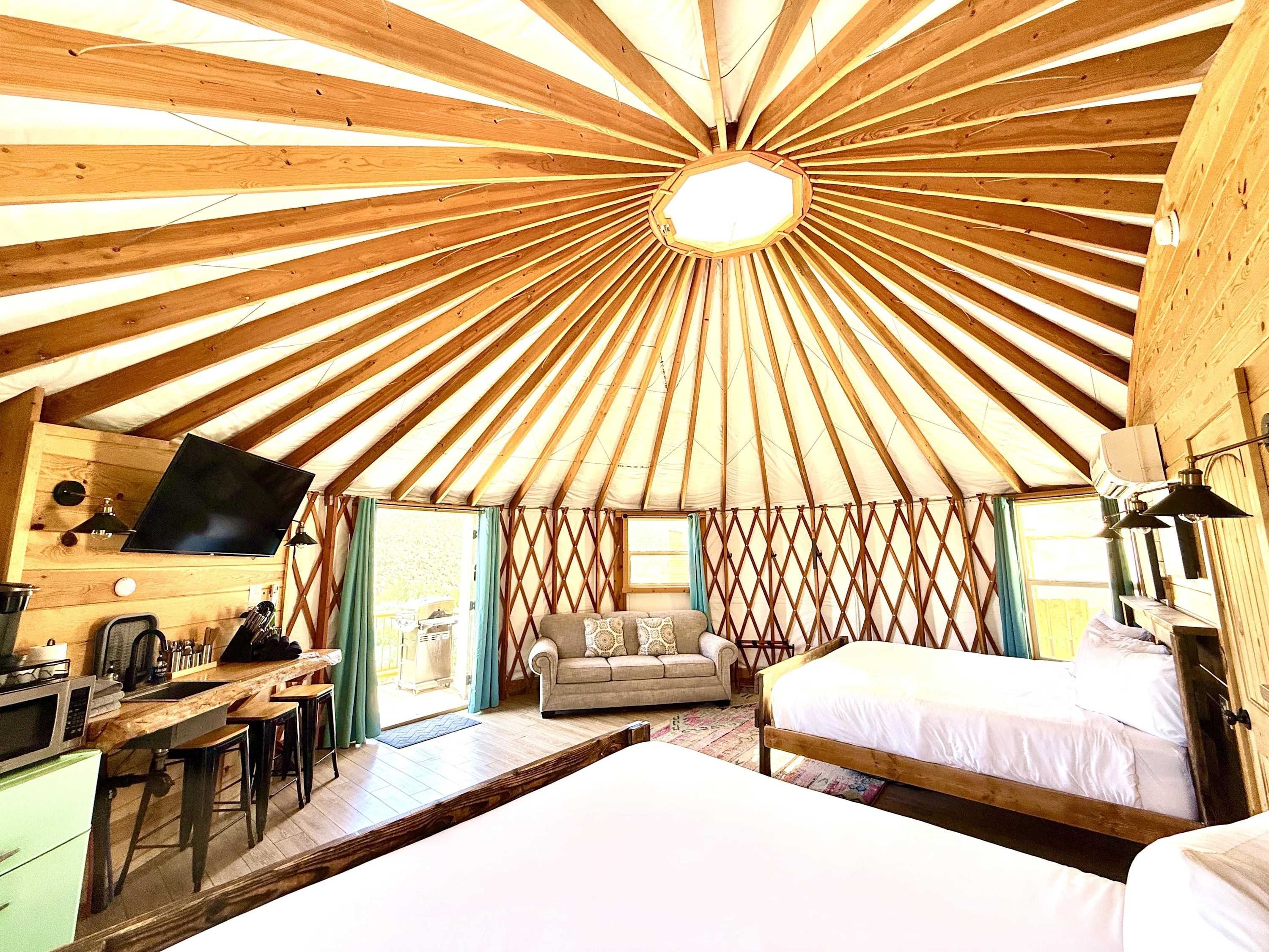 A yurt interior with a central dome, a sofa, a bed, a desk with a computer, and a kitchen area.