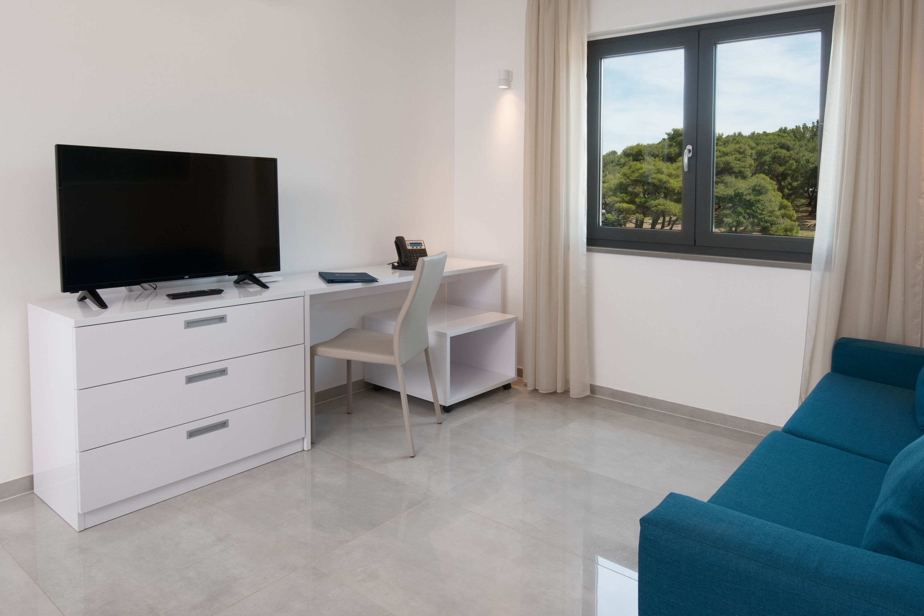Une chambre moderne équipée d’un téléviseur à écran plat, d’un bureau avec une chaise, d’un canapé bleu et d’une fenêtre donnant sur des arbres.