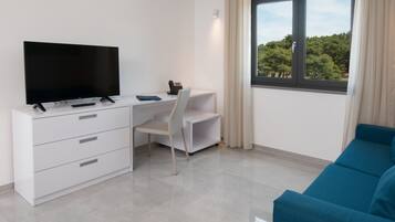 Une chambre moderne équipée d’un téléviseur à écran plat, d’un bureau avec une chaise, d’un canapé bleu et d’une fenêtre donnant sur des arbres.