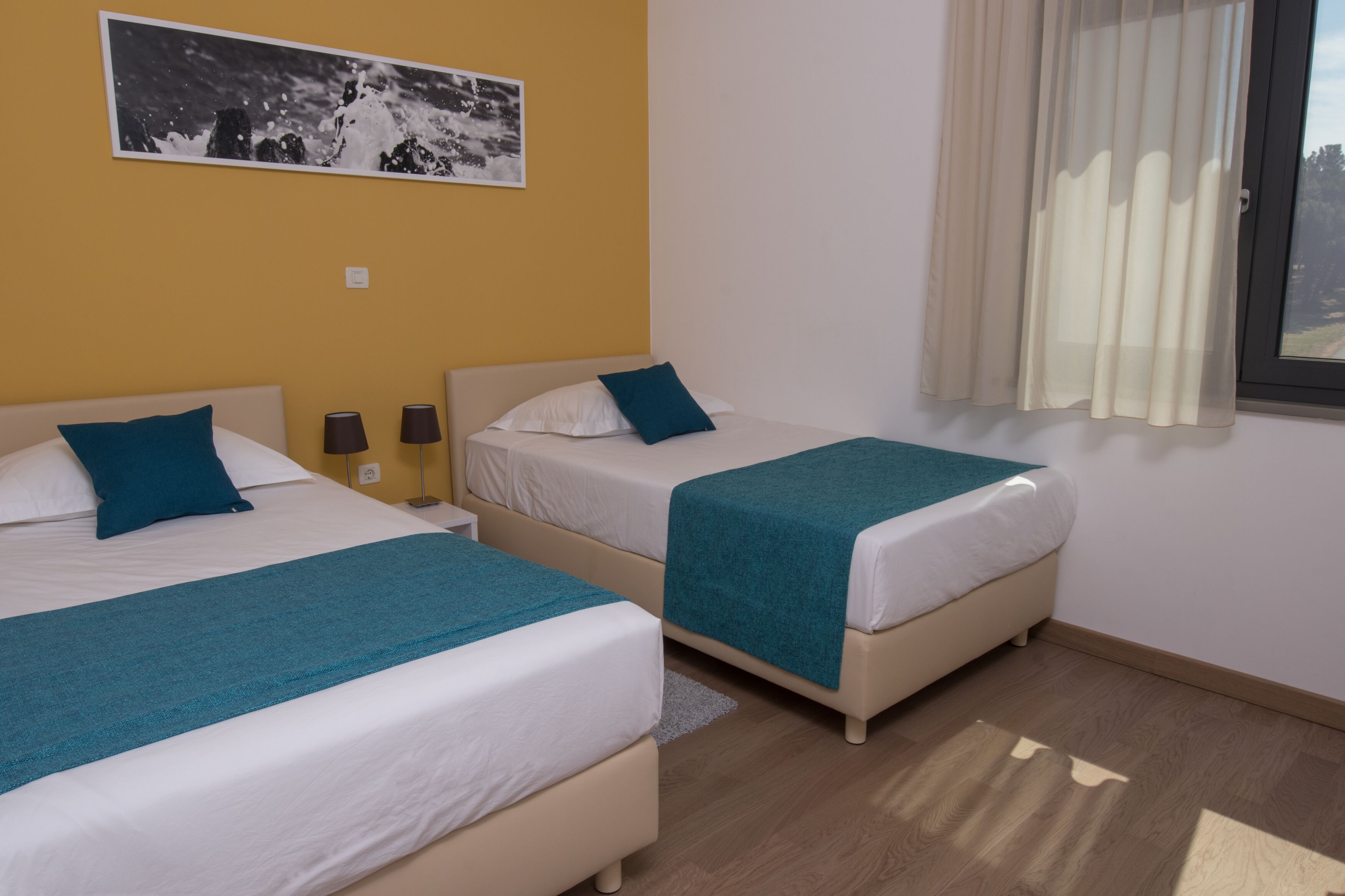 Habitació amb dos llits, cadascun amb matalàs blanc i roba de llit color aquamarina. Hi ha una finestra amb cortines translúcides, un quadre penjat a la paret i una petita catifa al terra.