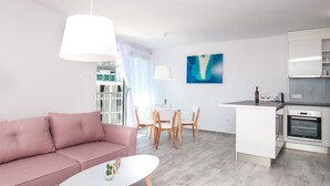 A modern living room with a pink sofa, a white coffee table, and a dining area with wooden chairs. There is a kitchen with white cabinets and a built-in oven. - Apartments Dva Galeba (Bol)