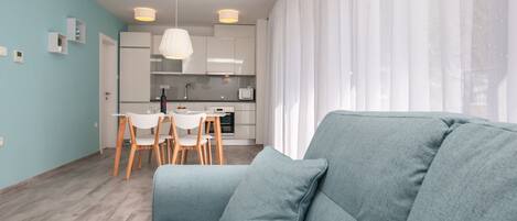 Uma sala de estar moderna com um sofá cinzento, uma área de refeições com cadeiras brancas e uma cozinha com armários brancos e uma luminária no teto branca.