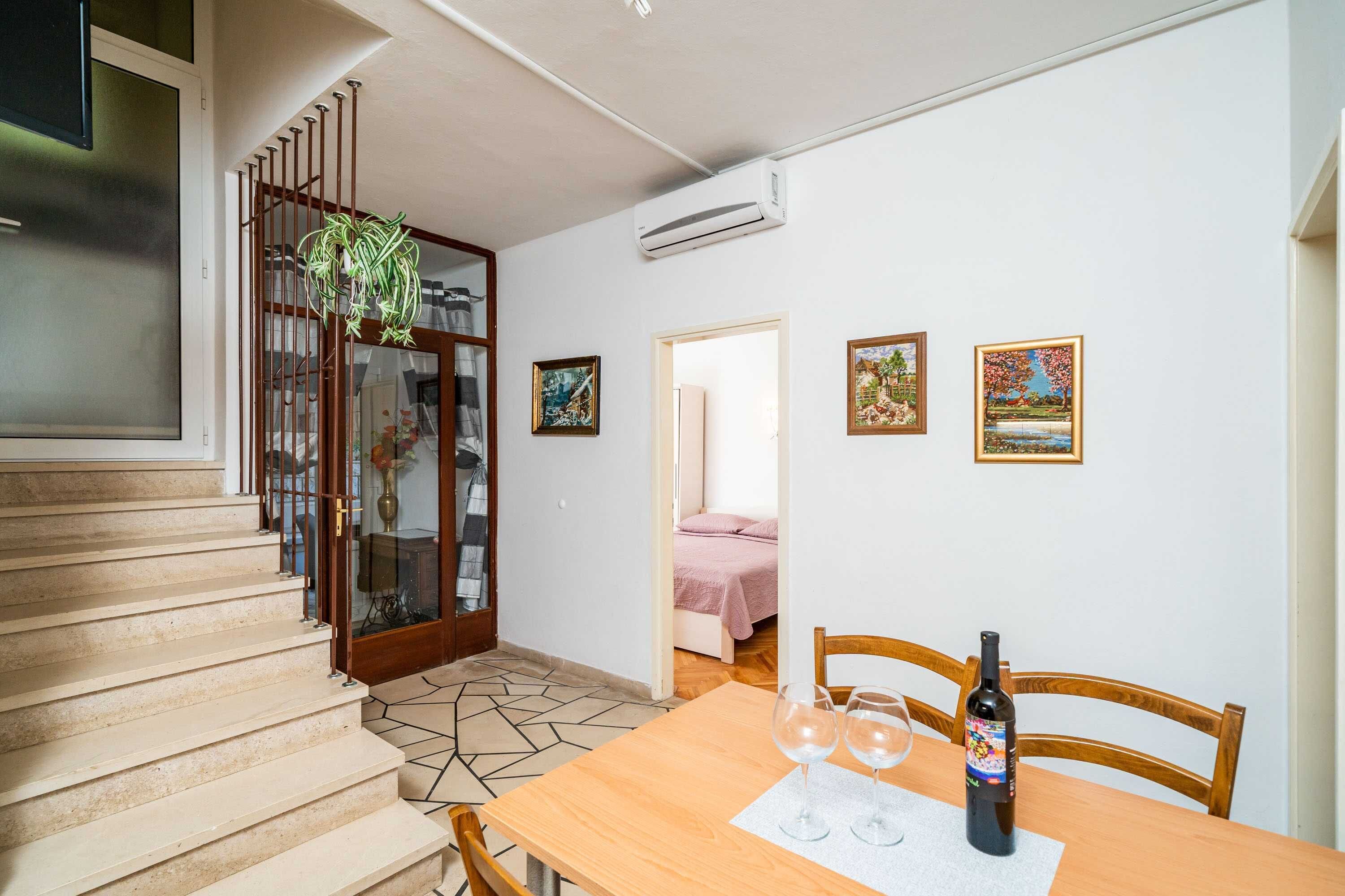 A dining area with a wooden table, two wine glasses, and a bottle of wine. There is a staircase with a glass door leading to another room.