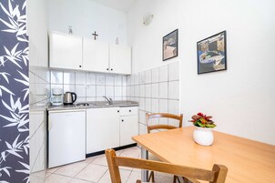 Una cocina compacta con armarios blancos, cafetera y lavamanos. Hay una mesa de comedor con sillas y un florero con flores. Dos cuadros están colgados en la pared.
