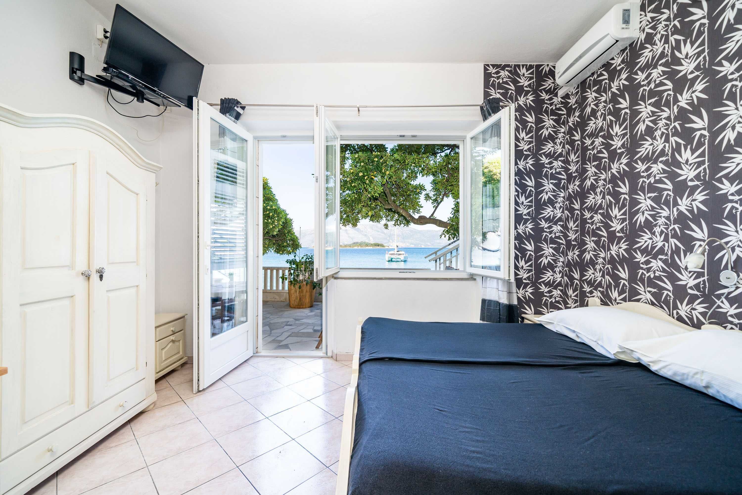 Quarto com cama, televisão presa à parede e vista para o mar por uma porta aberta.