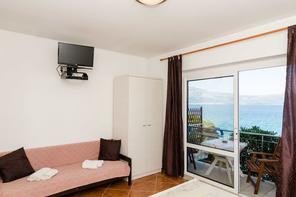 Een kamer met een roze bank, een aan de muur bevestigde televisie, een kledingkast en een balkon met een tafel en stoelen.