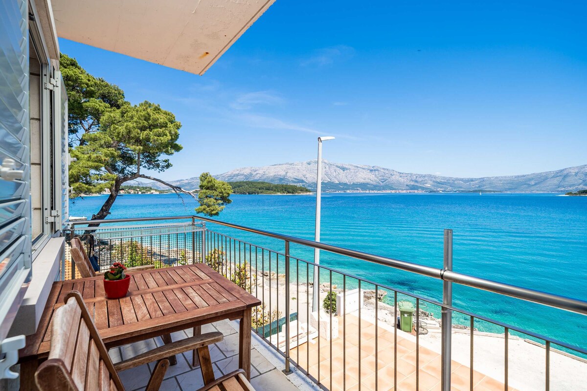 A balcony with a wooden table and chairs, overlooking a clear blue sea with mountains in the background.