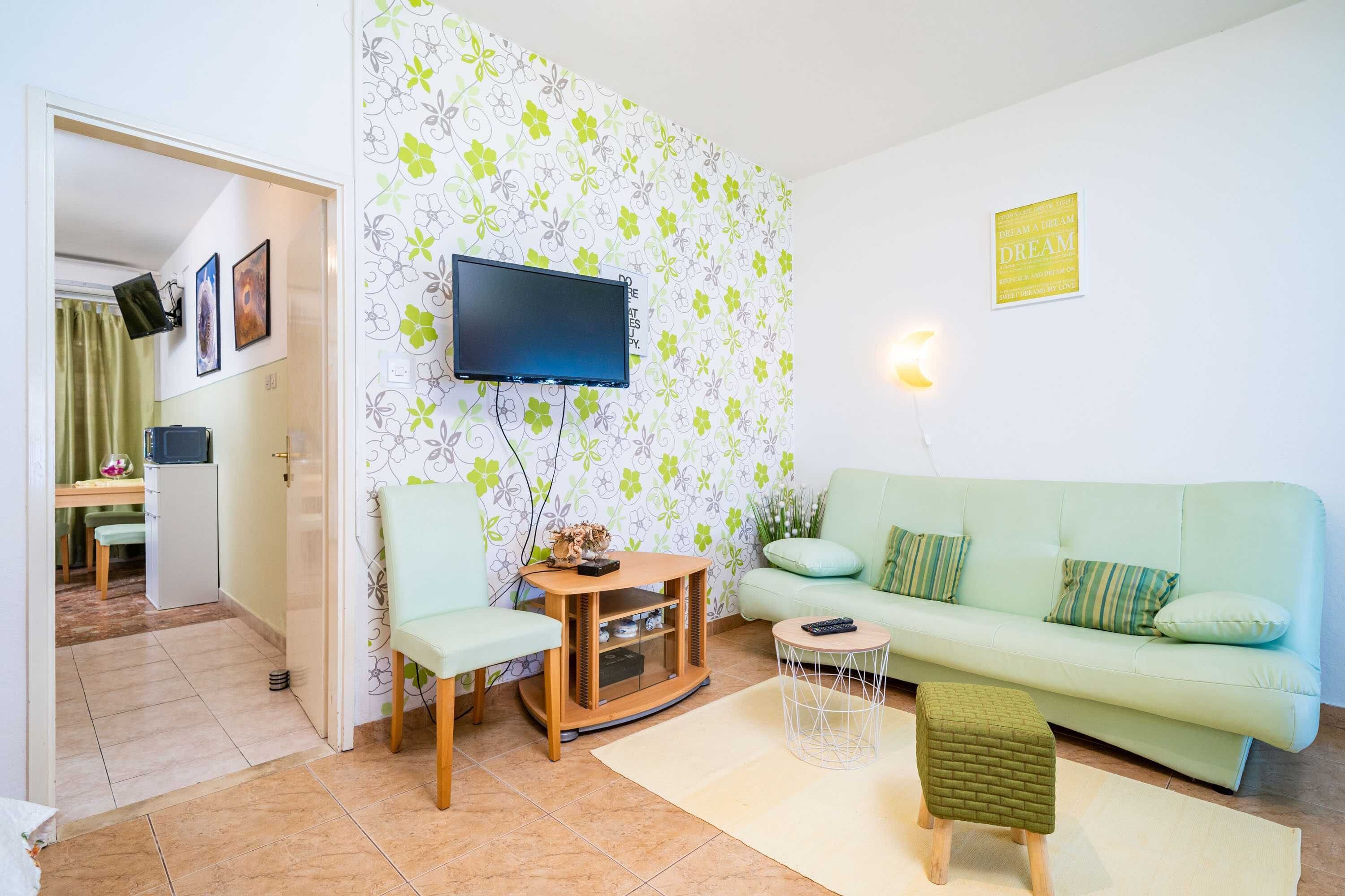 A living room with a flat-screen TV mounted on the wall, a light green sofa, a small wooden table, and a green ottoman.