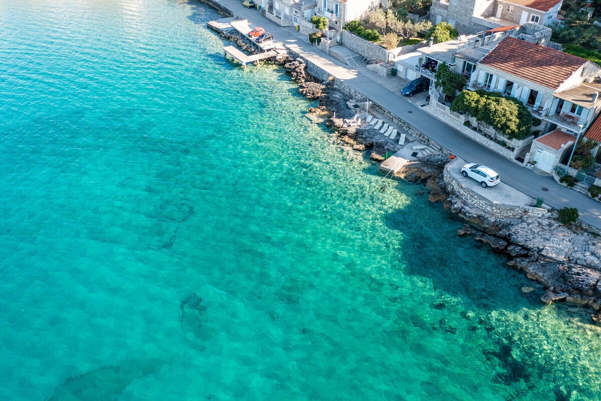 A coastal town with clear turquoise waters, stone buildings, and a paved walkway along the shore.
