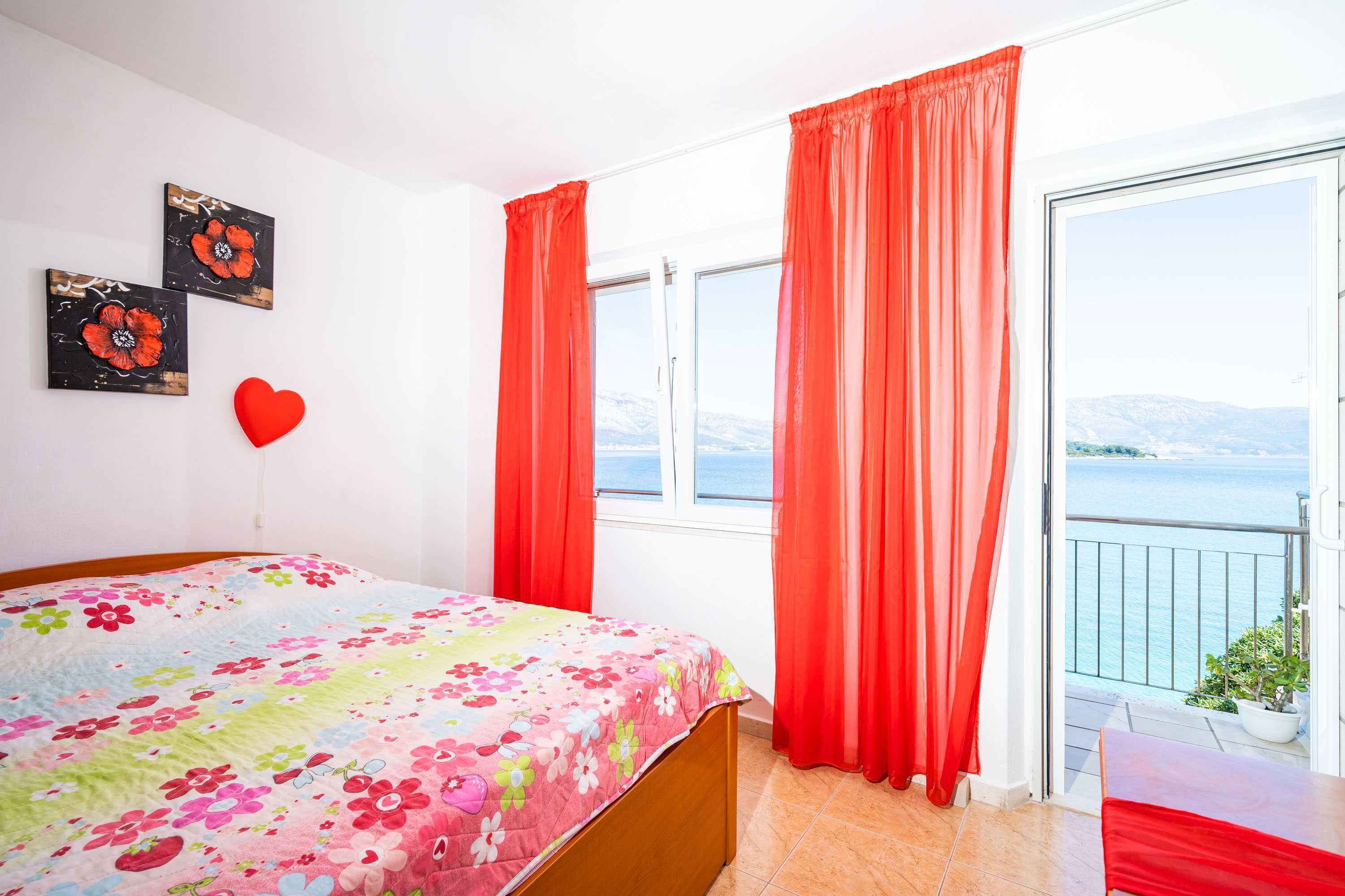 A bedroom with a bed, a window with curtains, a balcony, and a view of the sea.