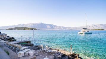 A clear view of a calm sea with a sailboat anchored, a rocky shore with lounge chairs, and a mountainous backdrop.