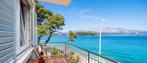 A balcony with a wooden table and chairs, overlooking a clear blue sea and a snow-capped mountain in the distance.