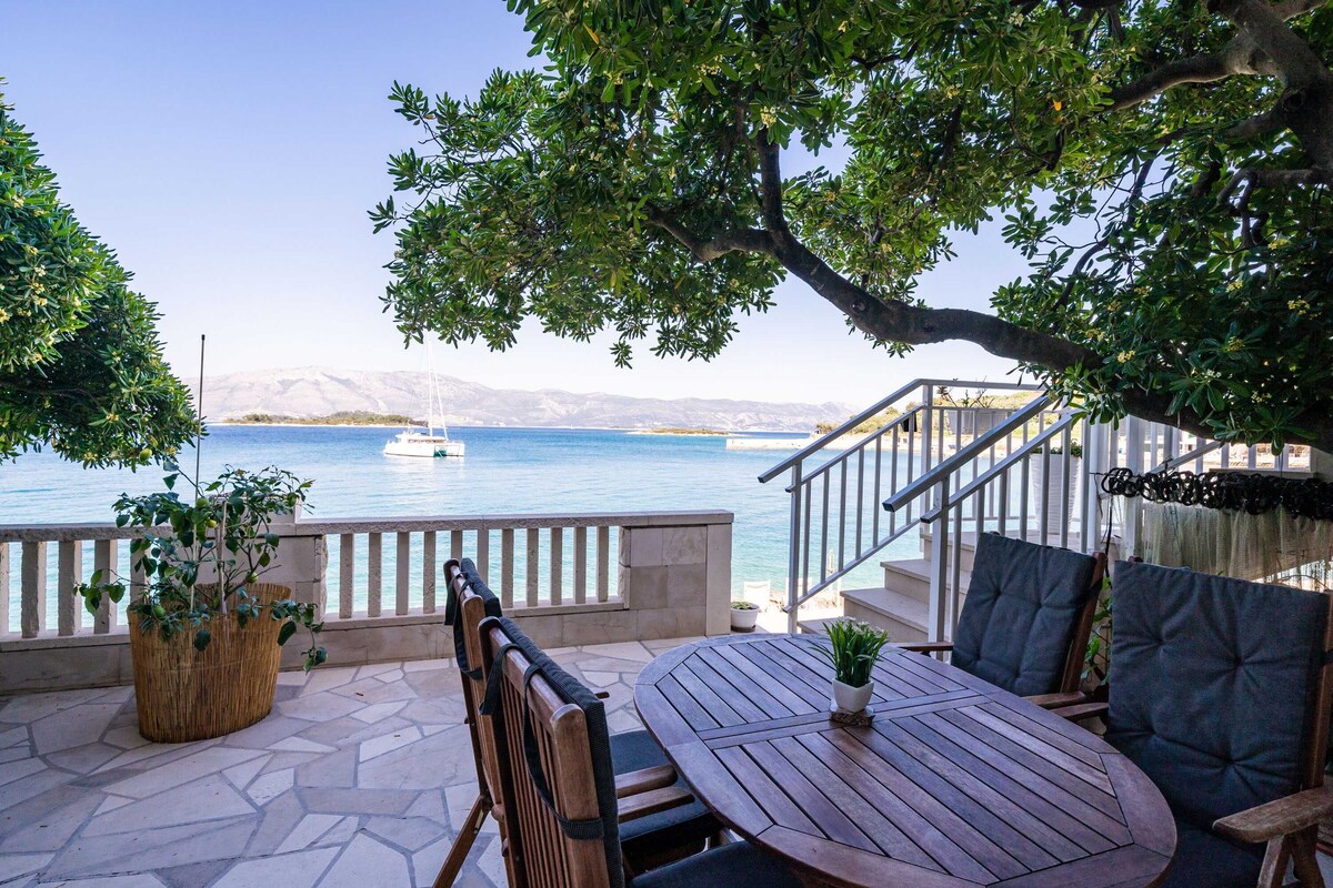 A wooden table with chairs, a potted plant, and a view of the sea with a boat.