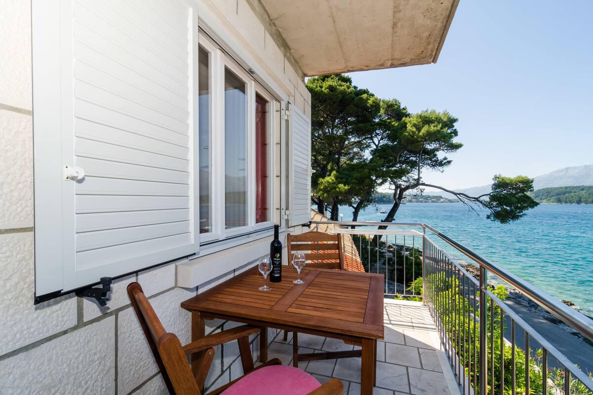 Een houten tafel met stoelen op een balkon, met uitzicht op zee en bomen.