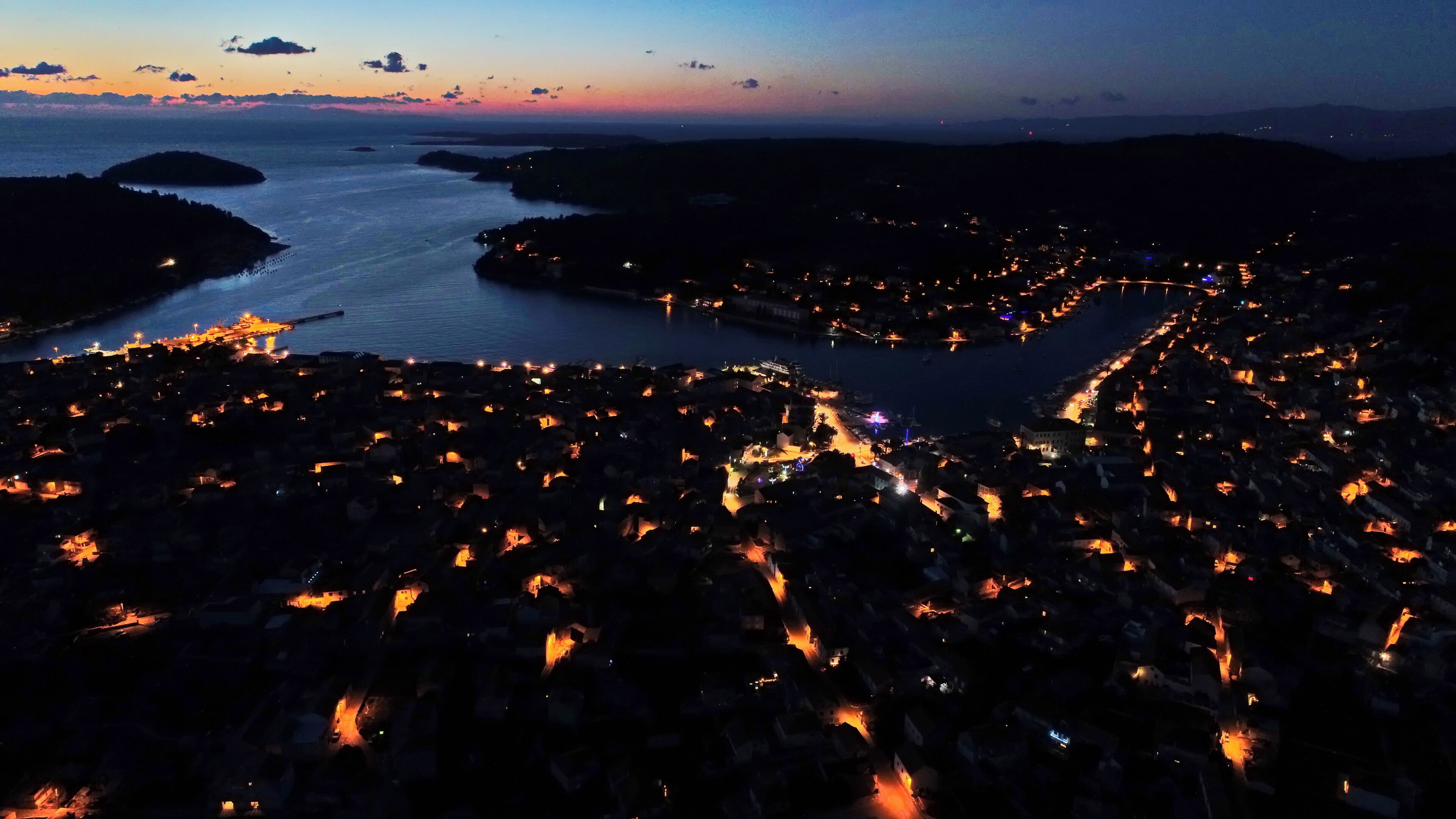 An aerial view of a coastal town at dusk with illuminated streets and buildings.