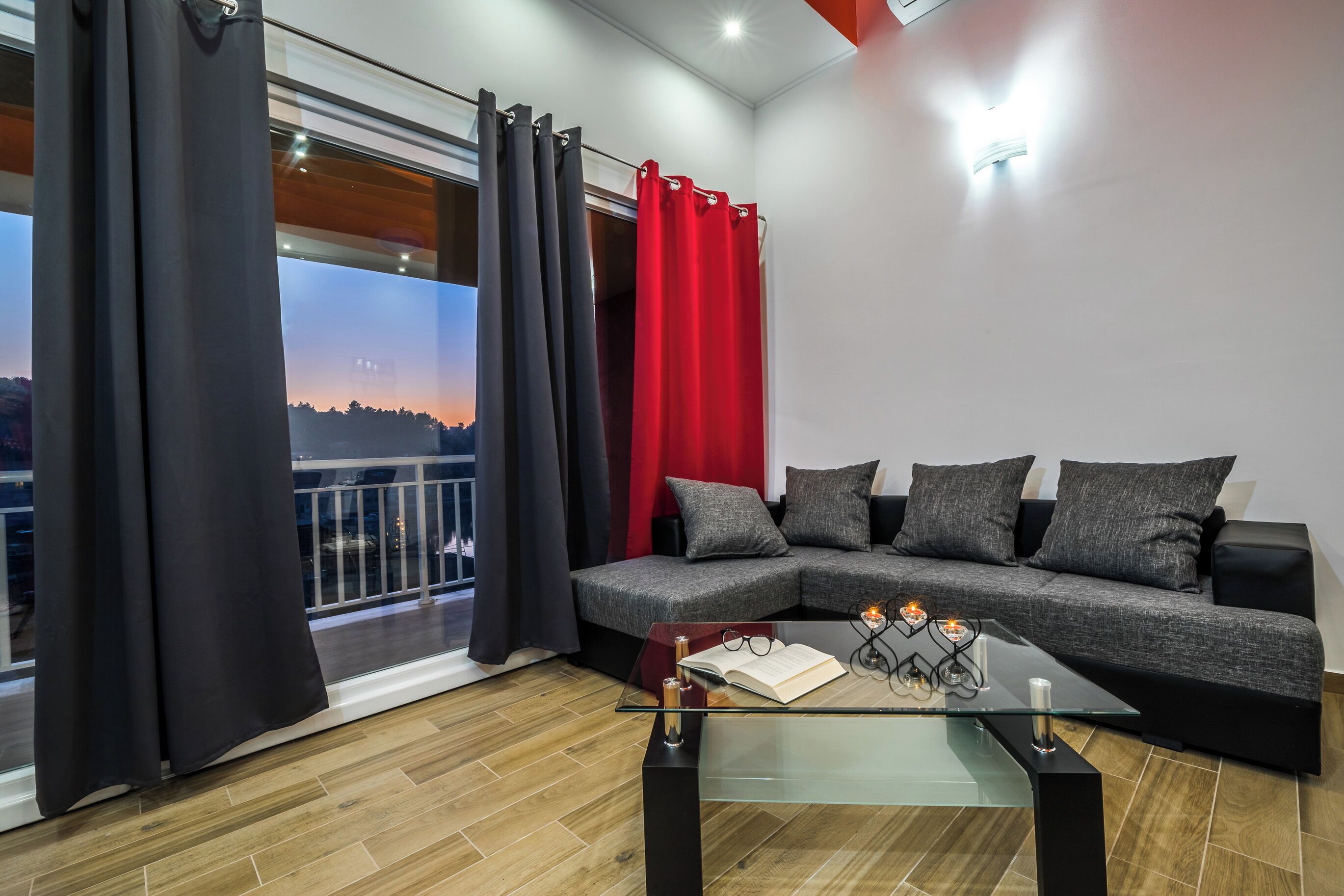 A modern living room with a glass coffee table, a grey sectional sofa, and black and red curtains.