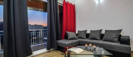 A modern living room with a glass coffee table, a grey sectional sofa, and black and red curtains.