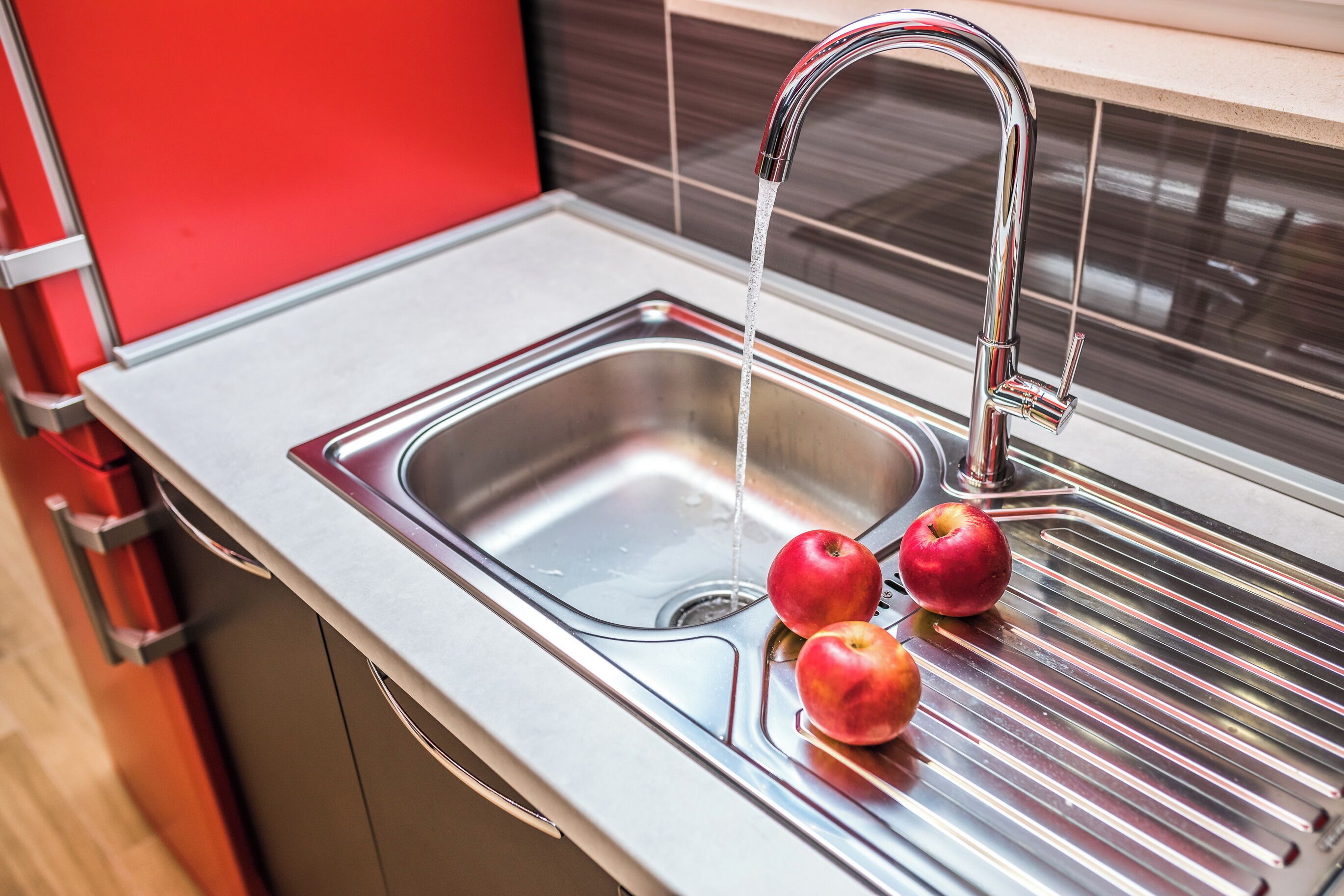 A stainless steel kitchen sink with a modern faucet, two apples, and a red cabinet.