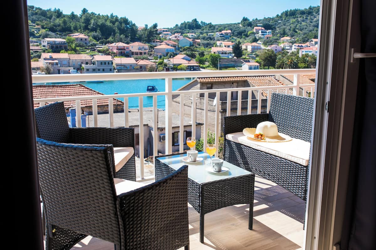 Ein Balkon mit Korbmöbeln, einem Tisch und Blick auf ein Schwimmbad und Häuser am Hang.