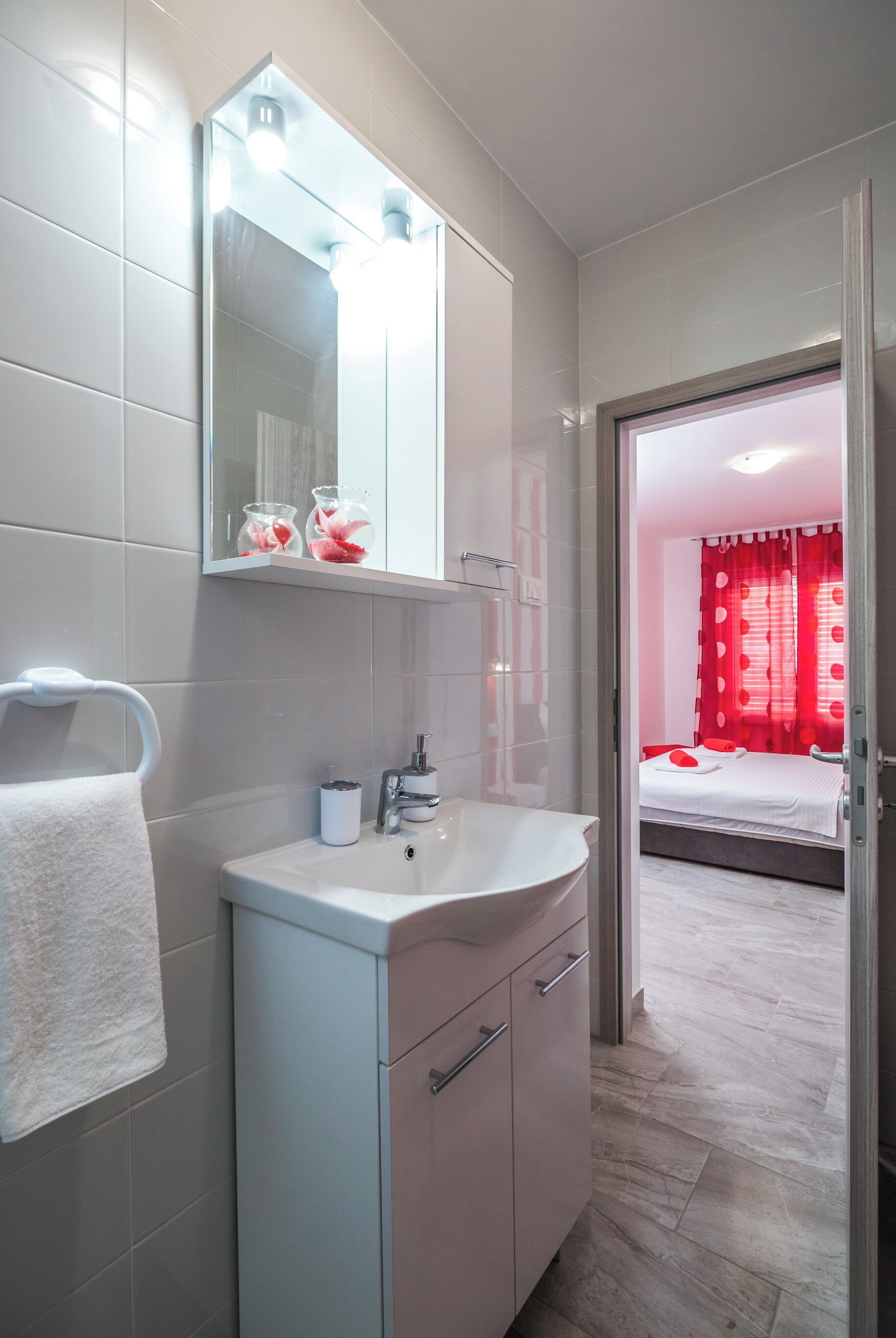 Une salle de bain moderne avec un lavabo blanc, un miroir et un porte-serviettes. Une chambre à coucher avec un lit et des rideaux rouges est visible à travers une porte ouverte.