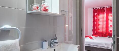 Une salle de bain moderne avec un lavabo blanc, un miroir et un porte-serviettes. Une chambre à coucher avec un lit et des rideaux rouges est visible à travers une porte ouverte.