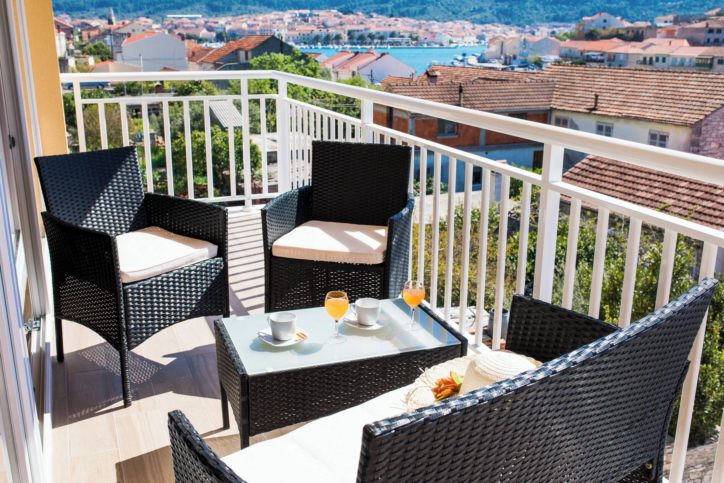 A balcony with wicker furniture, a table with two cups and two glasses, and a view of a town with red-roofed buildings.
