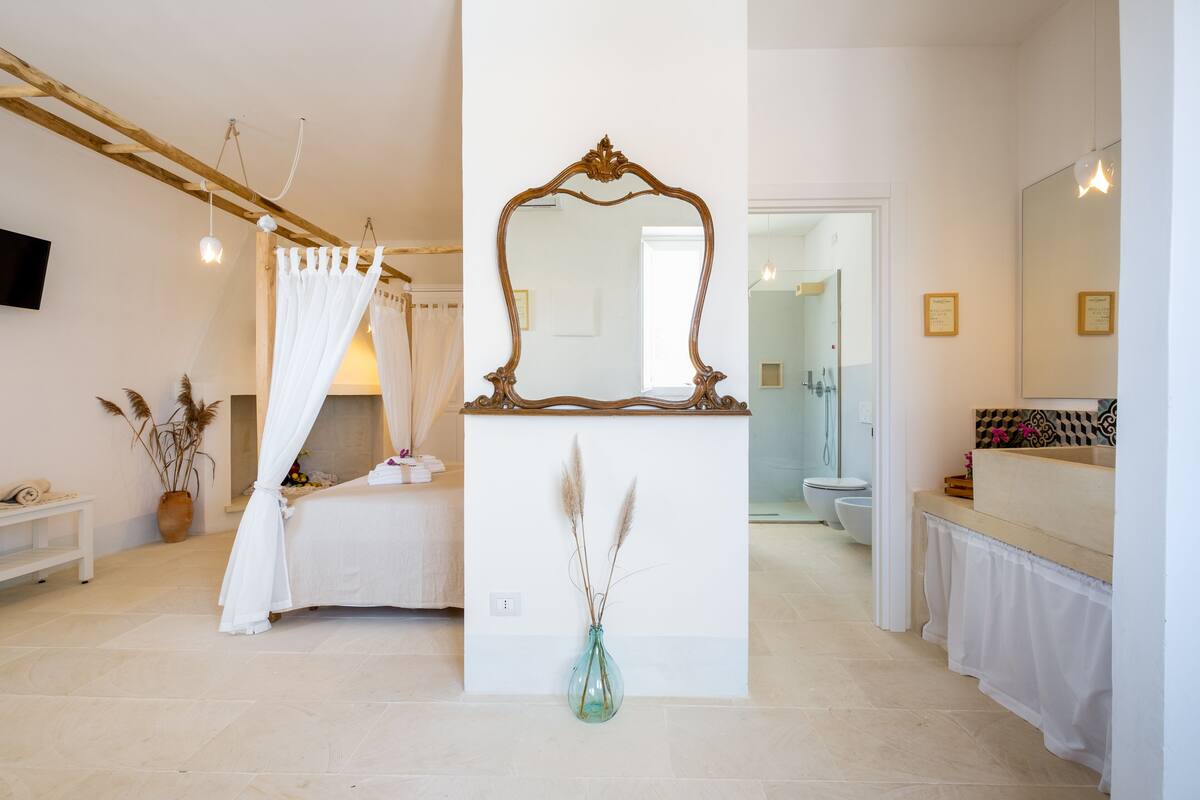 Ein Schlafzimmer mit Baldachinbett, einem großen Spiegel und einer Vase mit Trockenpflanzen.
