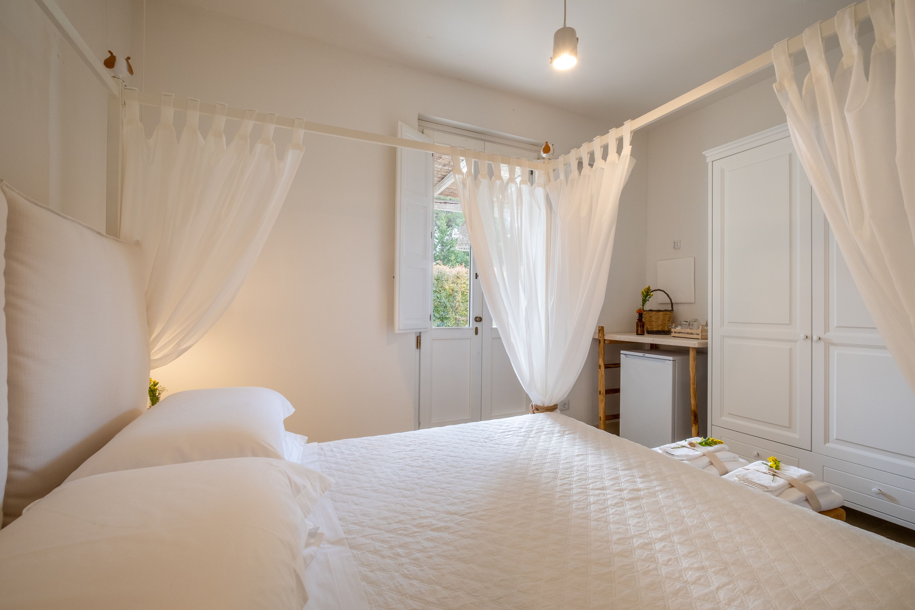 A bedroom with a large bed, white curtains, a small bedside table with a lamp, and a wardrobe.