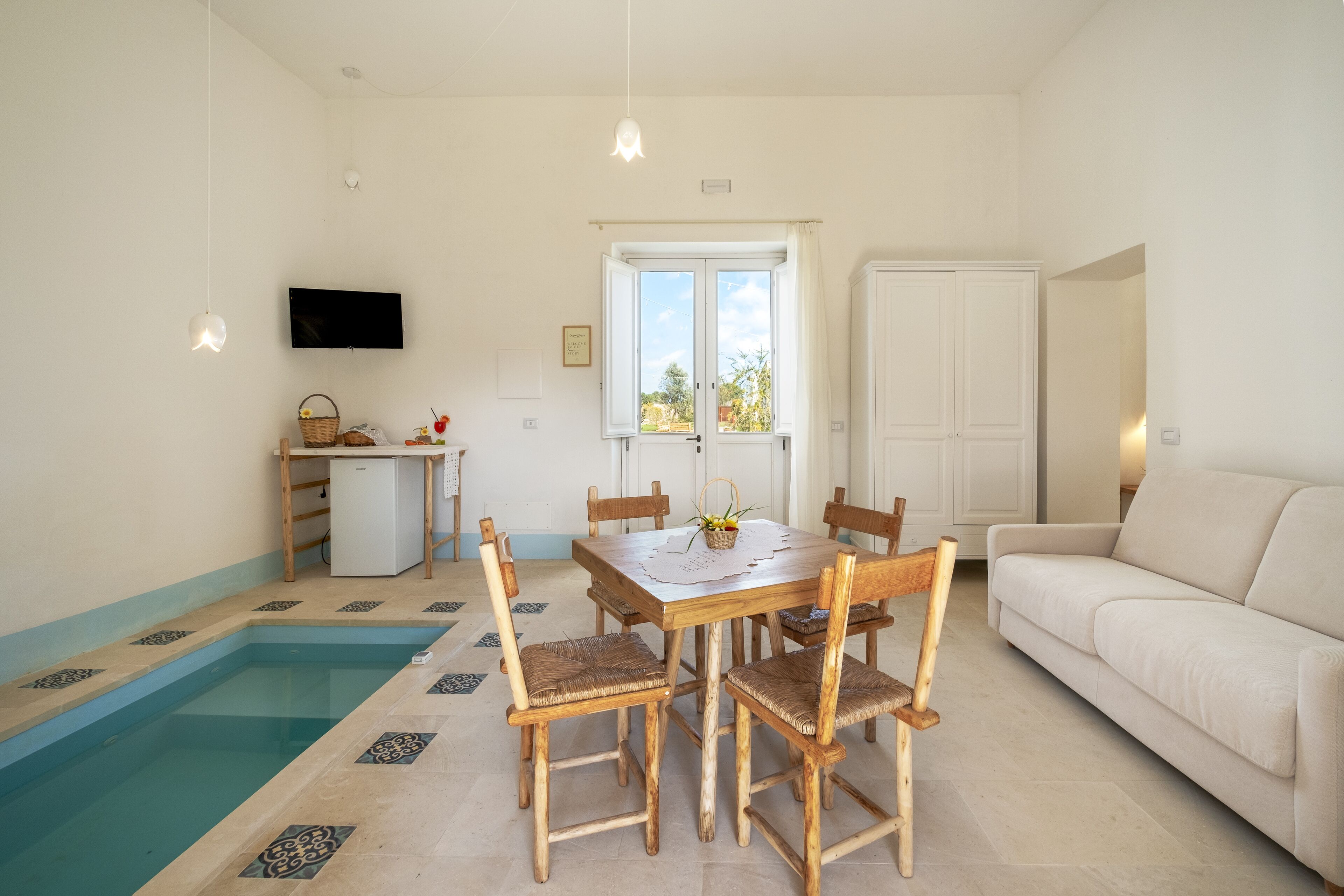 A room with a pool, a dining table with chairs, a sofa, a TV, and a window with curtains.