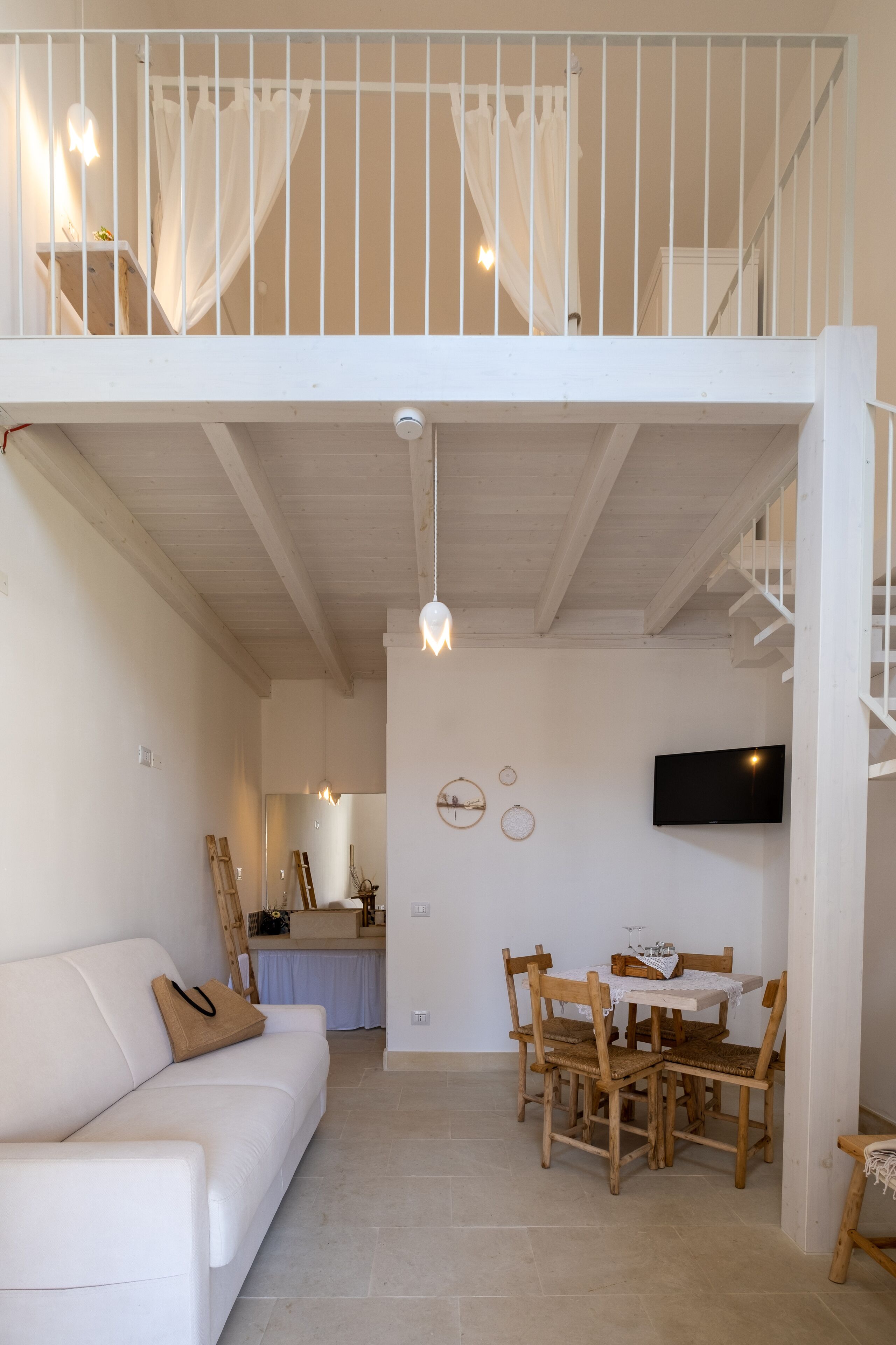 A spacious living area with a white sofa, wooden dining table, and chairs. A TV is mounted on the wall, and a staircase leads to an upper level.