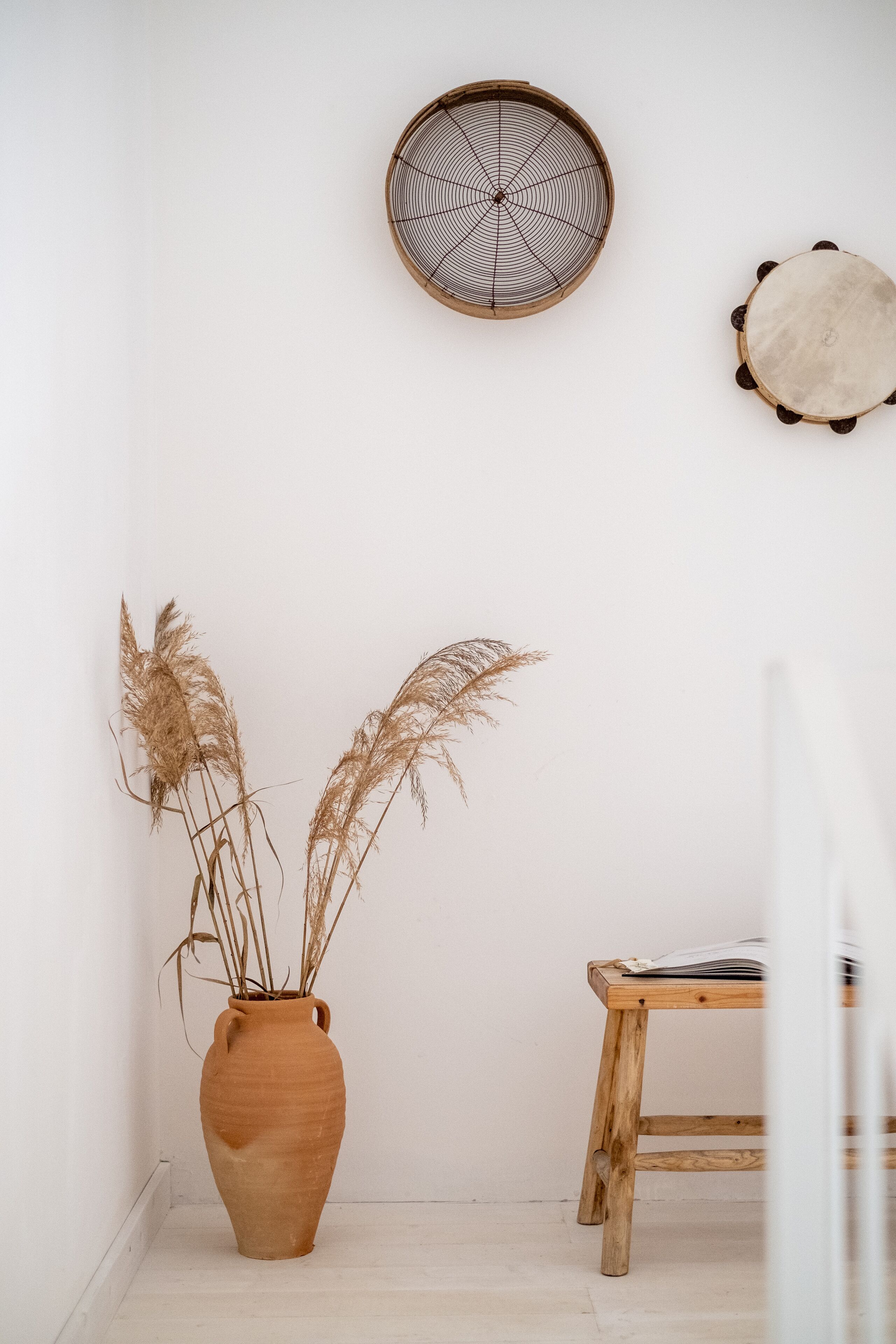 A room with a wooden stool, a terracotta vase with dried pampas grass, and a woven wall hanging.