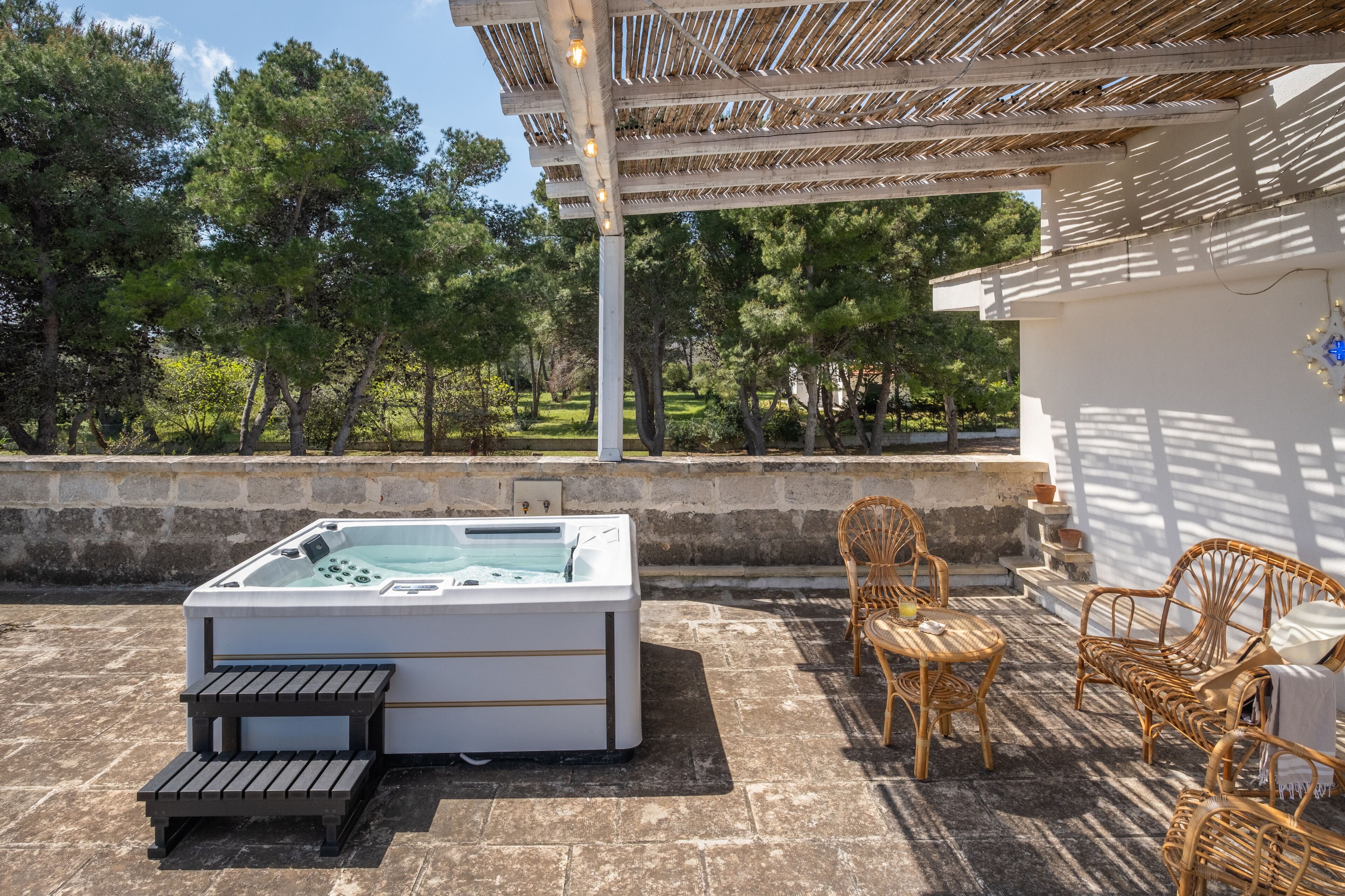 A hot tub with a control panel, a wooden bench, wicker chairs, and a pergola with lights.