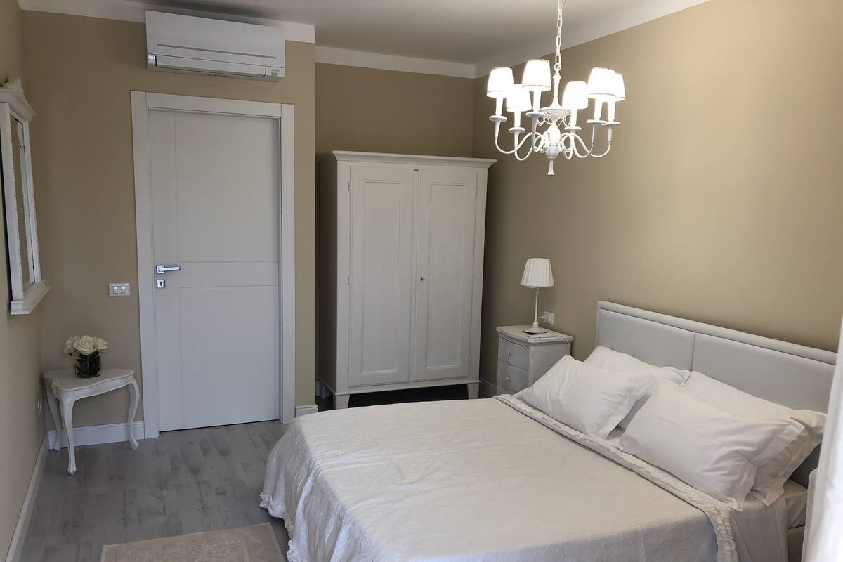 A bedroom with a bed, a nightstand, a lamp, a wardrobe, and an air conditioning unit.