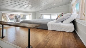 Een slaapkamer met een houten vloer, een bed met wit beddengoed, een ventilator en een raam met gordijnen.